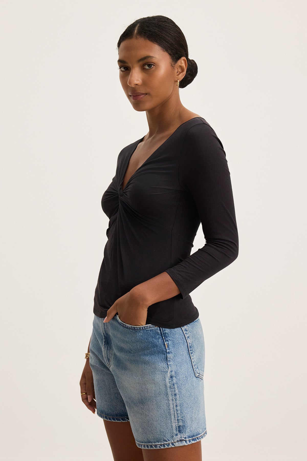 A person wearing the SETT TEE from Velvet by Graham & Spencer, a black, ultra-soft fabric top with a v-neckline, paired with denim shorts, stands with their hands in pockets against a plain background.