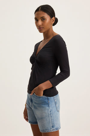 A person wearing the SETT TEE from Velvet by Graham & Spencer, a black, ultra-soft fabric top with a v-neckline, paired with denim shorts, stands with their hands in pockets against a plain background.