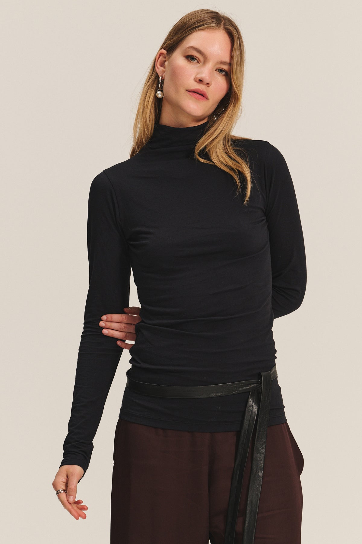 A woman in the TALISIA TEE by Velvet by Graham & Spencer—a black mock neck long-sleeve top—pairs it with brown pants and long earrings, standing against a plain light background for a timeless look.-40182502260929