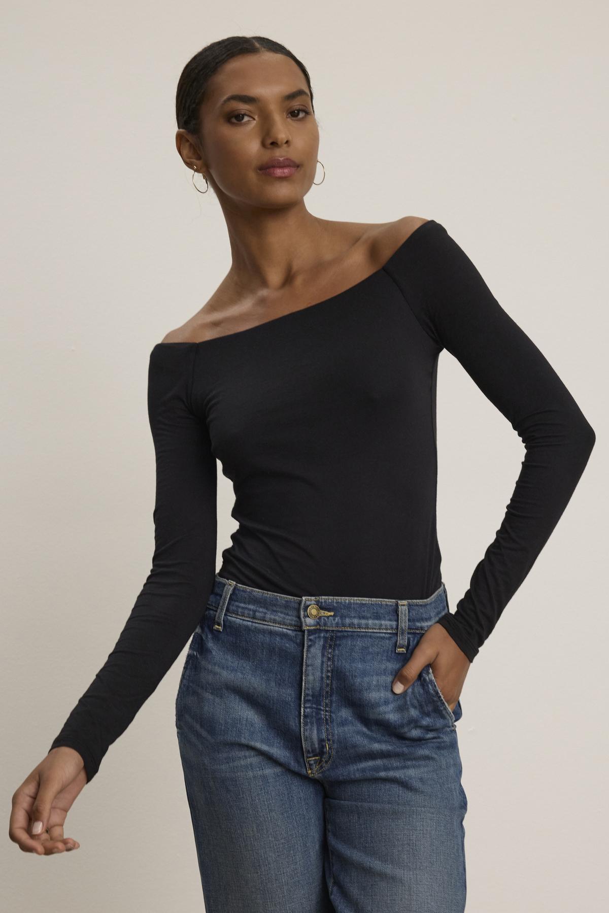 A person wearing the TANYA TEE, an off-the-shoulder black long-sleeve top by Velvet by Graham & Spencer, paired with blue jeans, stands against a plain background.