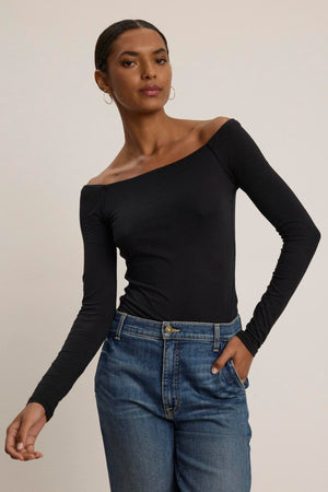 A person wearing the TANYA TEE, an off-the-shoulder black long-sleeve top by Velvet by Graham & Spencer, paired with blue jeans, stands against a plain background.