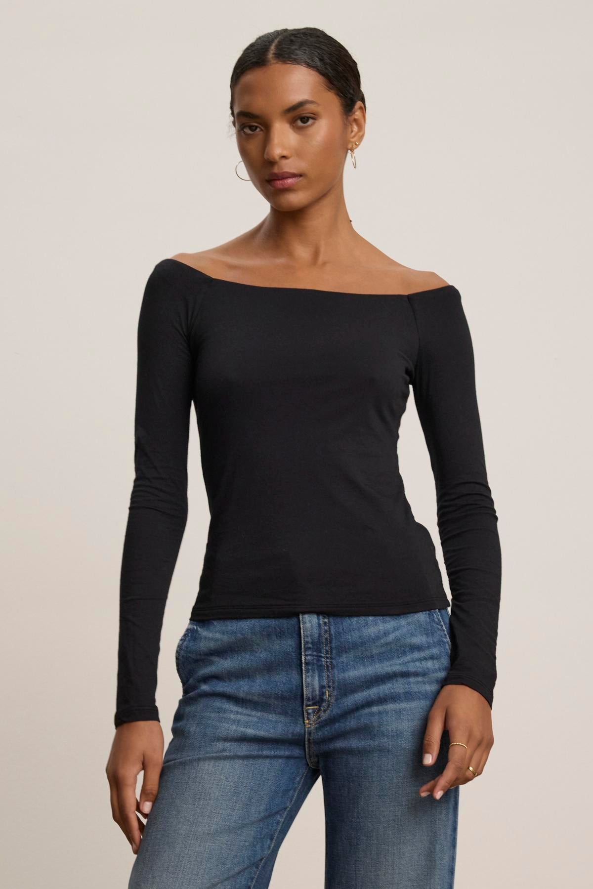 A person wearing the TANYA TEE, an off-the-shoulder top by Velvet by Graham & Spencer, along with jeans stands against a plain background.