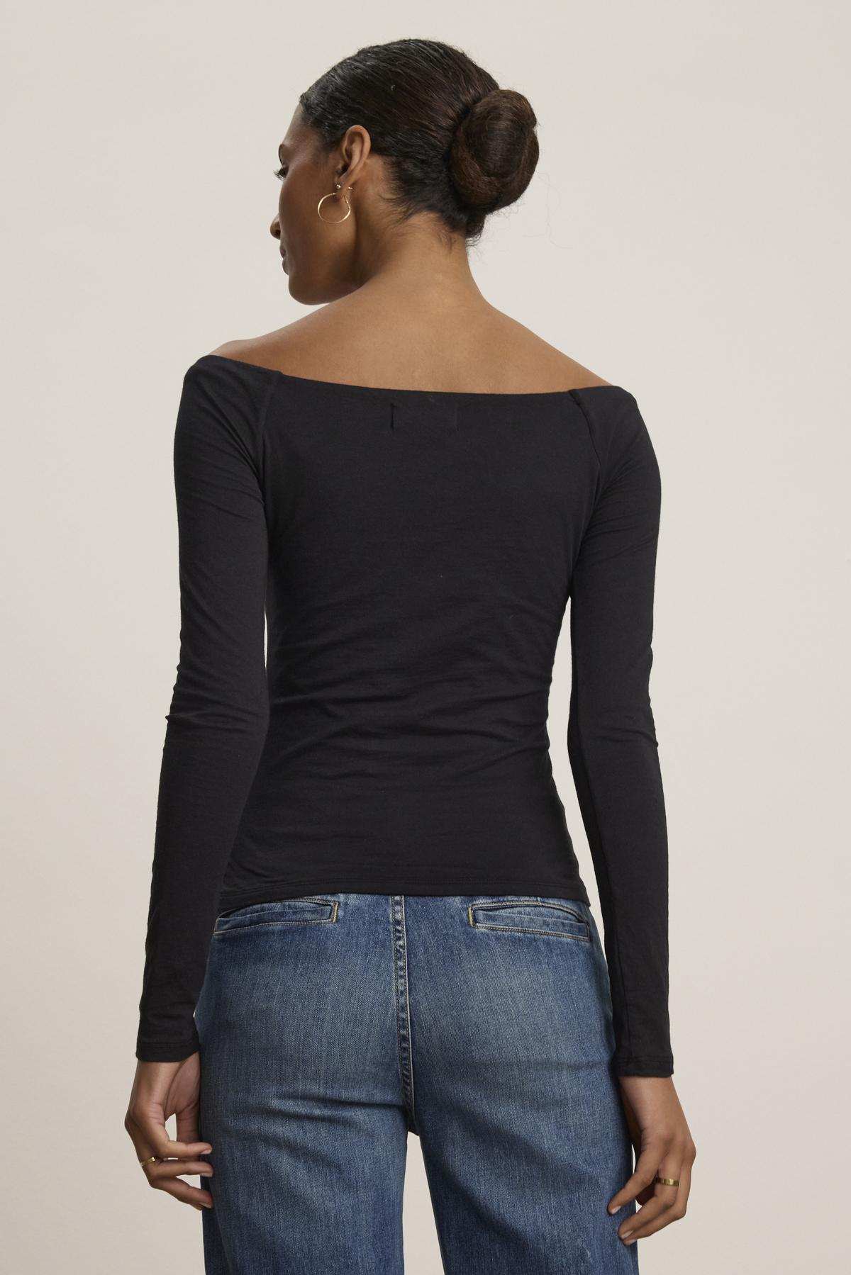 A person with tied-back hair is dressed in the TANYA TEE long-sleeve black top by Velvet by Graham & Spencer, paired with blue jeans, and facing away from the camera.