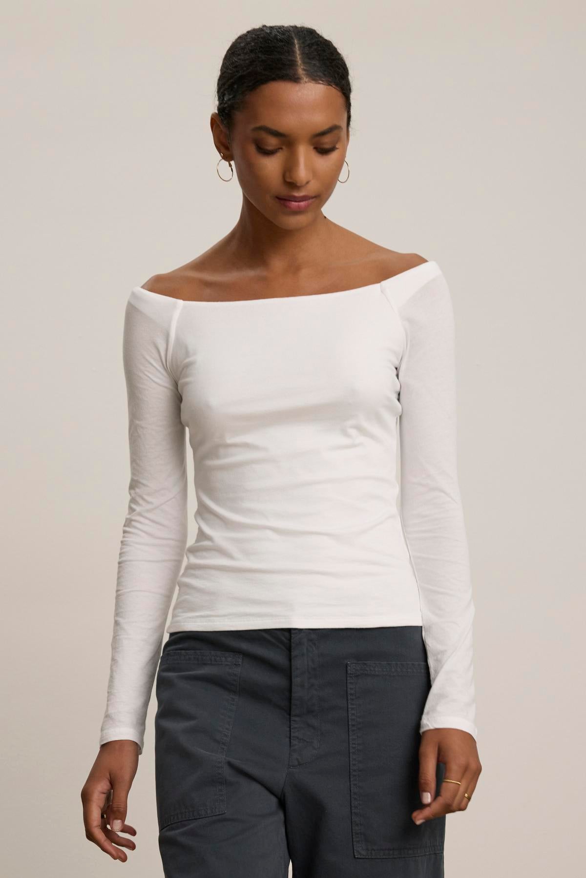 A person is standing against a plain background wearing the TANYA TEE, a white, long-sleeve, off-shoulder top by Velvet by Graham & Spencer, paired with gray pants.-38347958386881