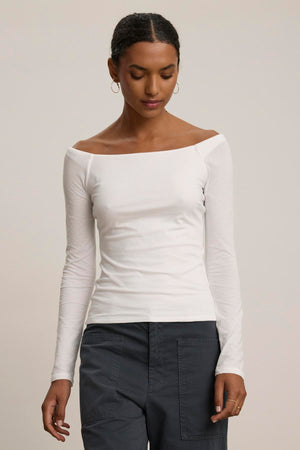 A person is standing against a plain background wearing the TANYA TEE, a white, long-sleeve, off-shoulder top by Velvet by Graham & Spencer, paired with gray pants.