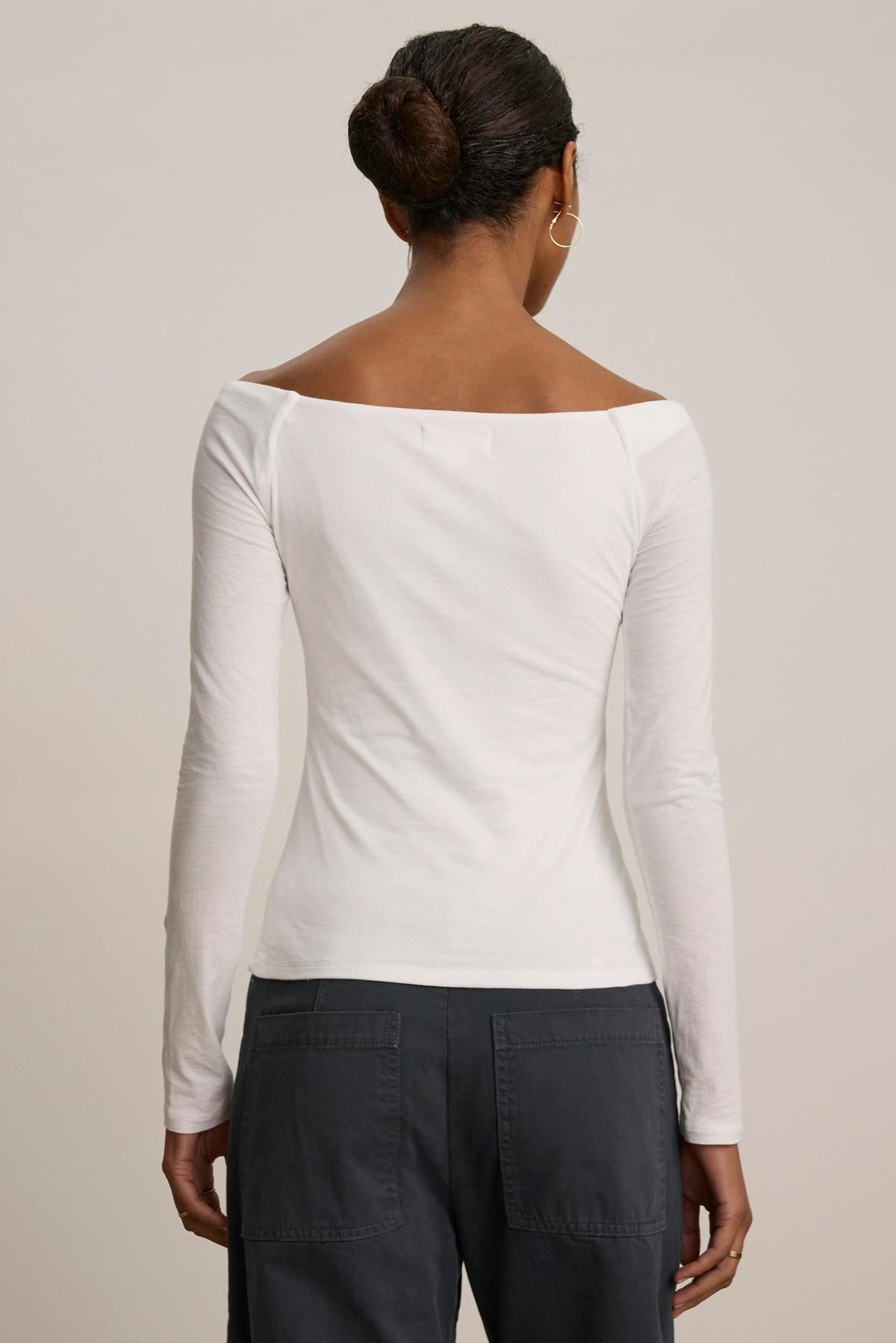A person wearing the TANYA TEE by Velvet by Graham & Spencer, an off-shoulder long-sleeve top in white, paired with dark pants, seen from the back against a neutral background.