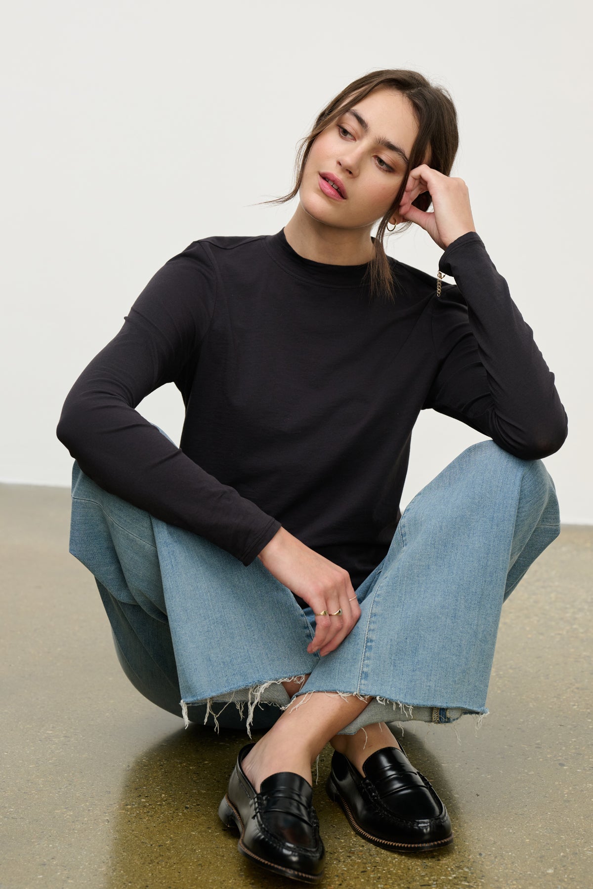 A woman wears the TRISH TEE by Velvet by Graham & Spencer with light blue frayed jeans, sitting on the floor with one arm on her knee and gazing to the side—capturing an effortless everyday look.-39814949339329