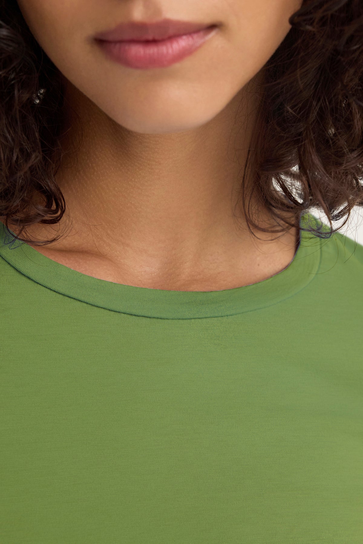 A curly-haired person is shown from the shoulders up, wearing Velvet by Graham & Spencer's ZOFINA TEE in green—a fitted long sleeve tee with a classic crew neckline. Their lips and chin are visible.-38928263839937