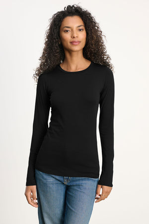 A person with curly hair wears the ZOFINA TEE by Velvet by Graham & Spencer—a fitted long sleeve crewneck—paired with blue jeans, standing against a neutral light background.