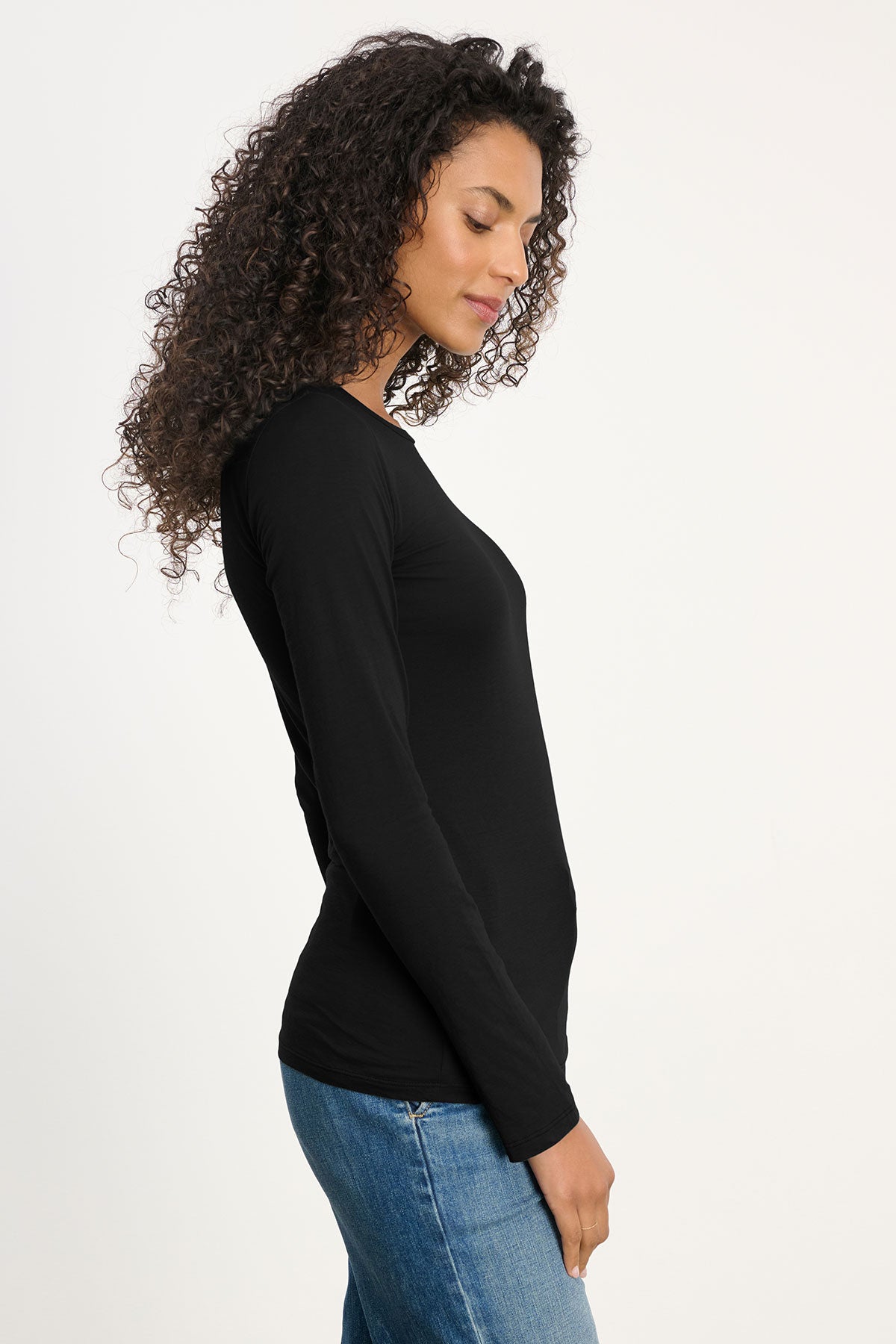 A person with long curly hair stands in profile against a plain light background, wearing the ZOFINA TEE by Velvet by Graham & Spencer—a fitted long sleeve top with a classic crew neckline—paired with blue jeans.