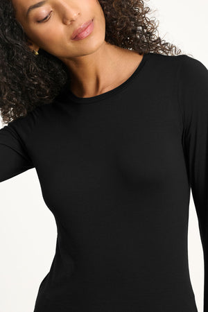 A woman with curly hair models the ZOFINA TEE by Velvet by Graham & Spencer—a fitted, ultra-soft long sleeve crewneck—shown from the shoulders up against a plain light background.