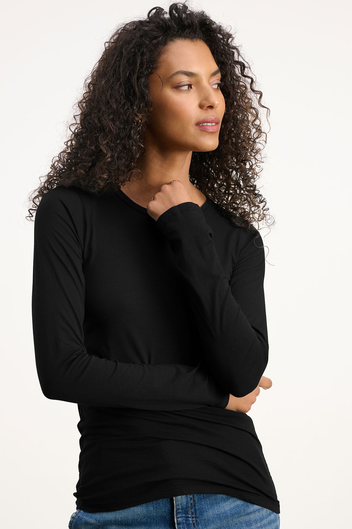 A woman with curly hair models the ZOFINA TEE by Velvet by Graham & Spencer in black, paired with blue jeans, standing against a plain light background and looking to the side with one hand raised near her chin.-39808287244481