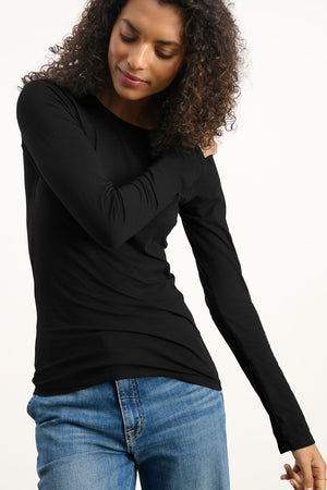 Wearing the ZOFINA TEE by Velvet by Graham & Spencer—a fitted long sleeve crew neck top—paired with blue jeans, the person stands against a plain light background.