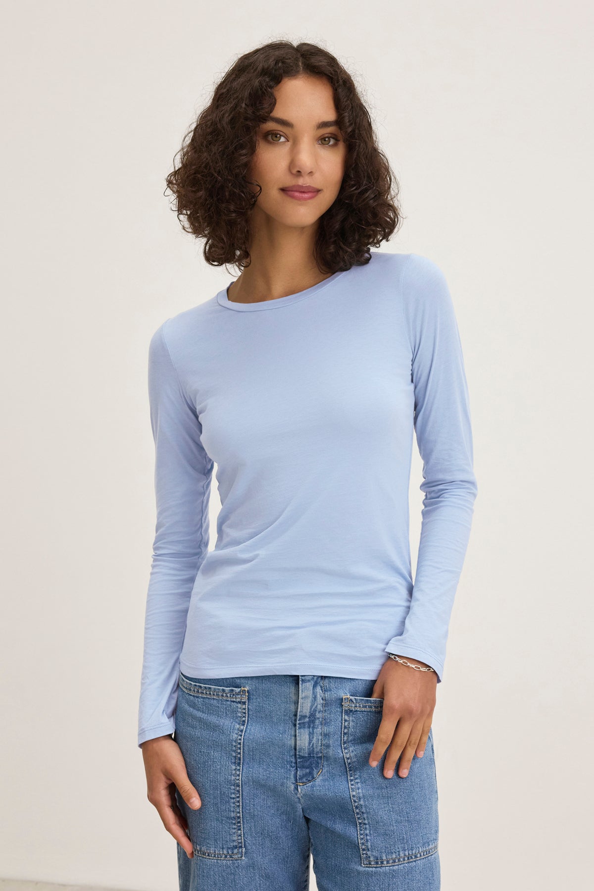 A person with curly hair wears a Velvet by Graham & Spencer ZOFINA TEE—an ultra-soft gauzy whisper light blue long-sleeve shirt—and blue jeans, standing against a plain background.-38928263872705
