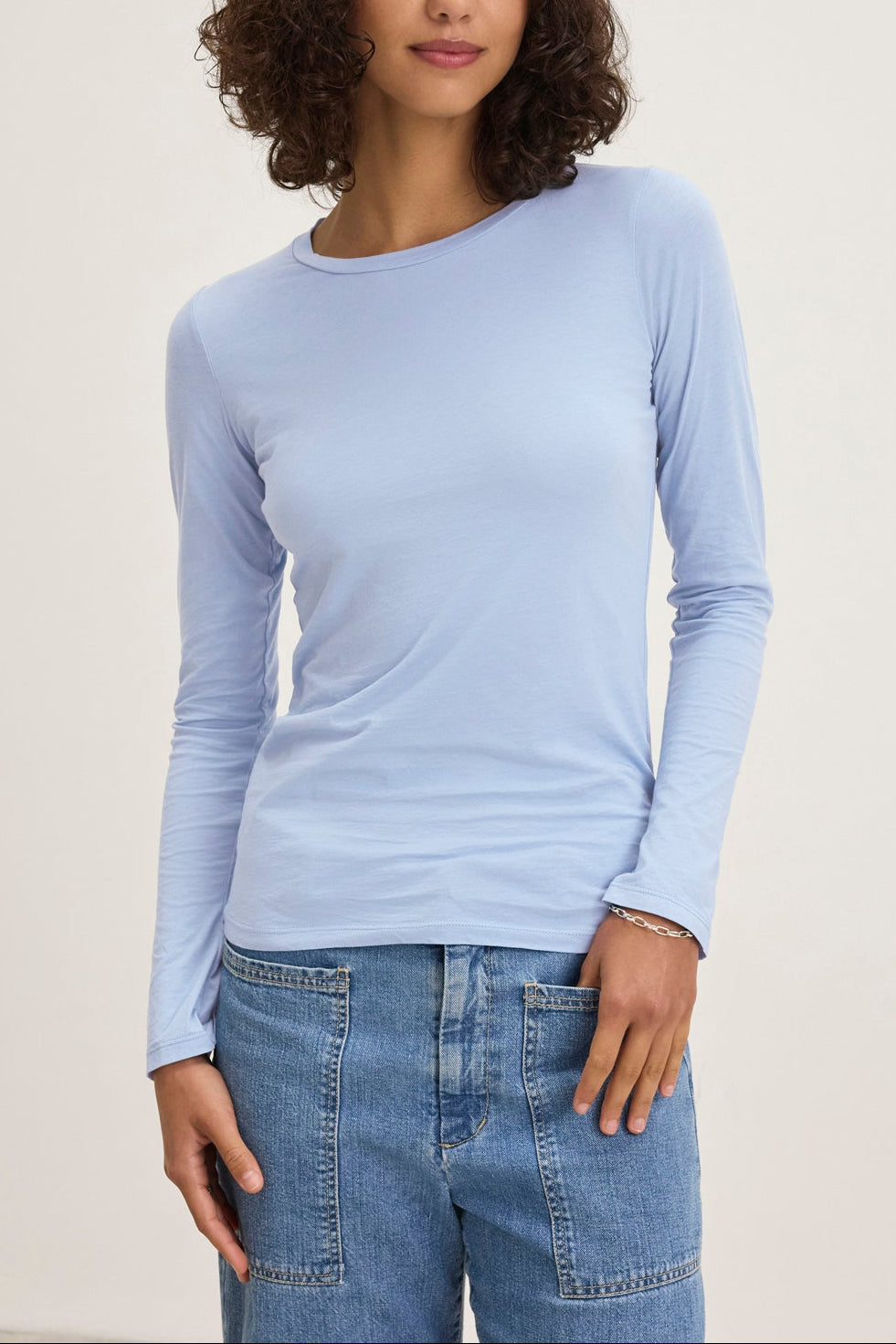 A person with curly hair wears a Velvet by Graham & Spencer ZOFINA TEE—an ultra-soft gauzy whisper light blue long-sleeve shirt—and blue jeans, standing against a plain background.