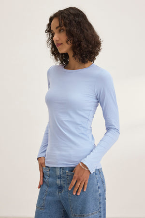 A person with curly hair, wearing the ZOFINA TEE by Velvet by Graham & Spencer in an ultra-soft gauzy whisper light blue fitted long sleeve style and blue jeans, stands against a plain background, casting a sideward glance.
