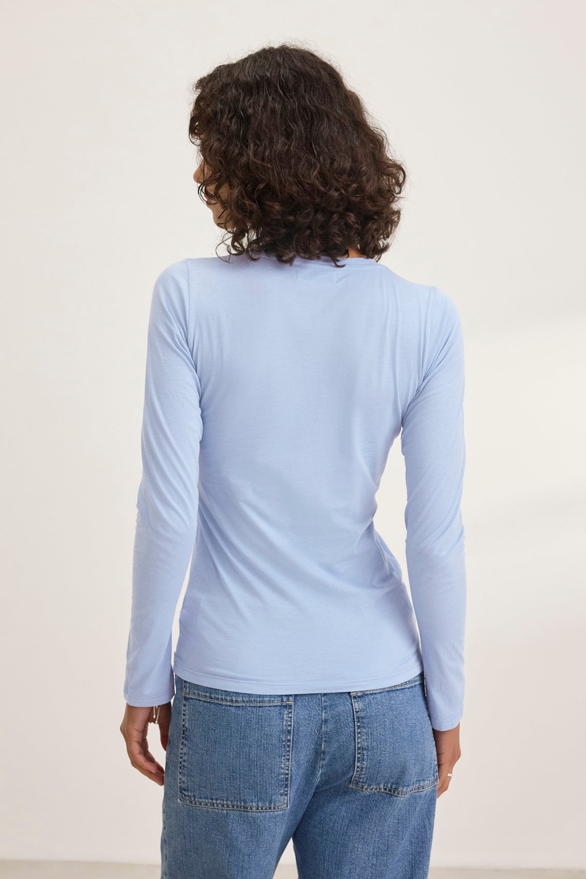 A person wearing the ZOFINA TEE in ultra-soft gauzy whisper light blue, featuring a classic crew neckline by Velvet by Graham & Spencer, and blue jeans stands facing away in a simple setting.-38928263938241