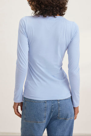 A person wearing the ZOFINA TEE in ultra-soft gauzy whisper light blue, featuring a classic crew neckline by Velvet by Graham & Spencer, and blue jeans stands facing away in a simple setting.