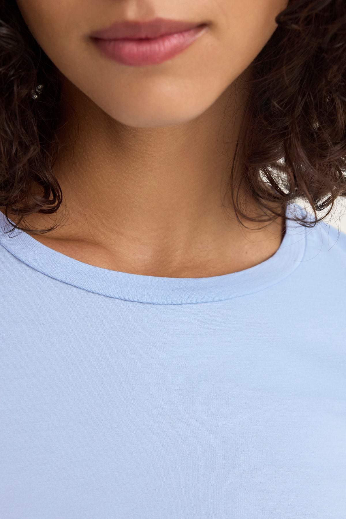 Close-up of a person wearing the ZOFINA TEE by Velvet by Graham & Spencer, featuring a light blue color and classic crew neckline, with the lower face and neck visible and curly hair cascading on the sides.-38928263971009