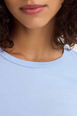 Close-up of a person wearing the ZOFINA TEE by Velvet by Graham & Spencer, featuring a light blue color and classic crew neckline, with the lower face and neck visible and curly hair cascading on the sides.