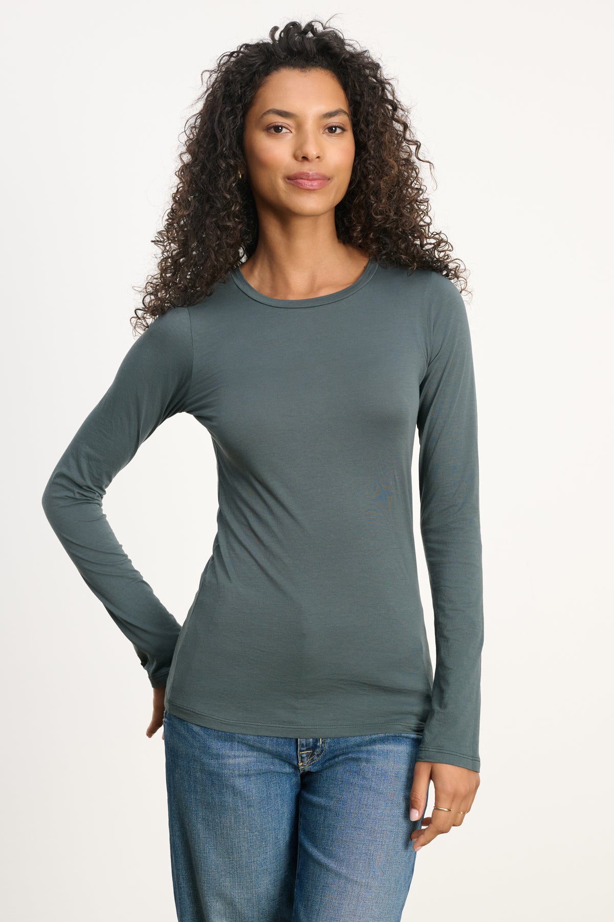 A woman with curly hair wears the ZOFINA TEE by Velvet by Graham & Spencer—an ultra-soft, fitted long sleeve crewneck in dark green—with blue jeans, standing against a plain light background.-39742401970369