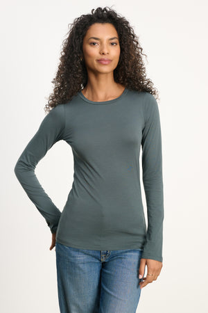 A woman with curly hair wears the ZOFINA TEE by Velvet by Graham & Spencer—an ultra-soft, fitted long sleeve crewneck in dark green—with blue jeans, standing against a plain light background.