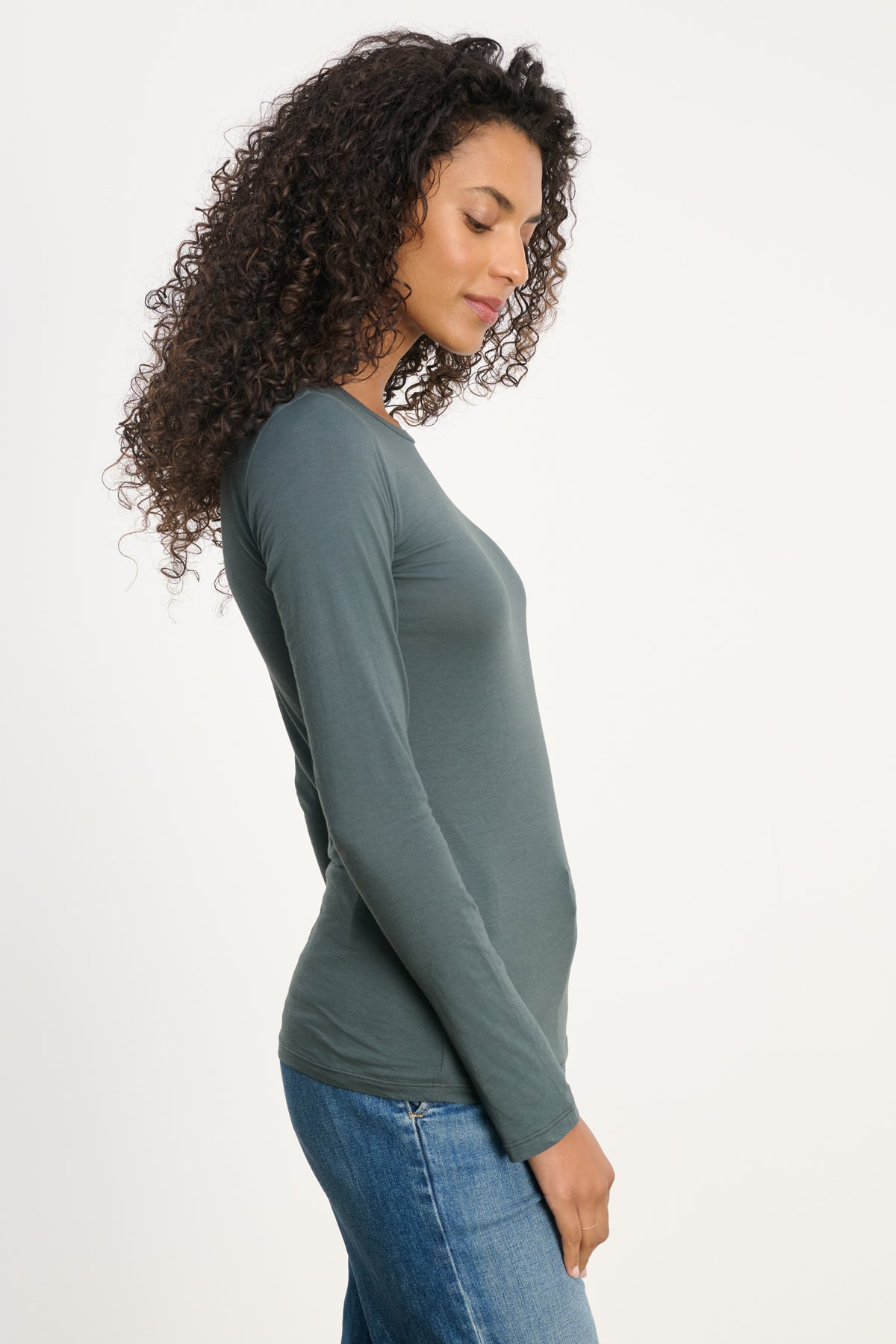 A person with long curly hair stands in profile, wearing the ultra-soft Velvet by Graham & Spencer ZOFINA TEE with a crew neckline and blue jeans against a plain white background.