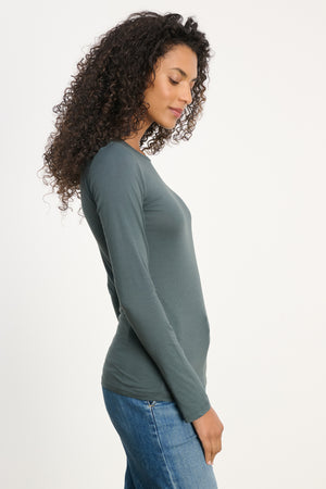 A person with long curly hair stands in profile, wearing the ultra-soft Velvet by Graham & Spencer ZOFINA TEE with a crew neckline and blue jeans against a plain white background.