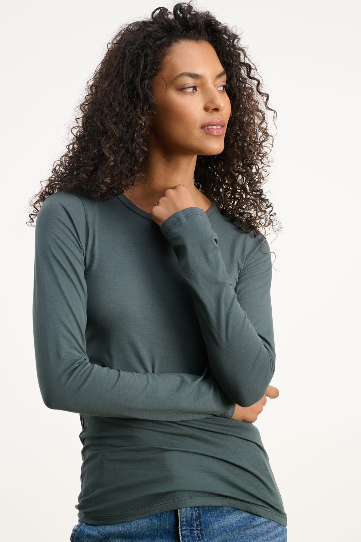 A woman with curly hair stands against a plain background, looking to the side, wearing the ZOFINA TEE by Velvet by Graham & Spencer—an ultra-soft, fitted dark green long sleeve crewneck—paired with blue jeans.