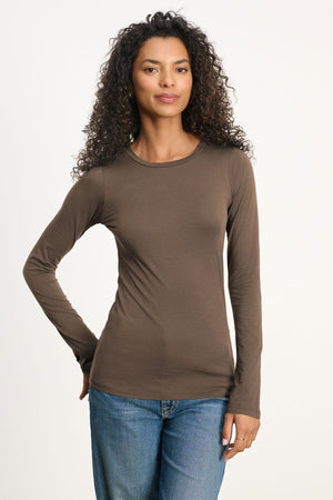 A person with long curly hair wears the ZOFINA TEE by Velvet by Graham & Spencer—an ultra-soft, fitted long sleeve top with a classic crew neckline—paired with blue jeans, standing against a plain light background.