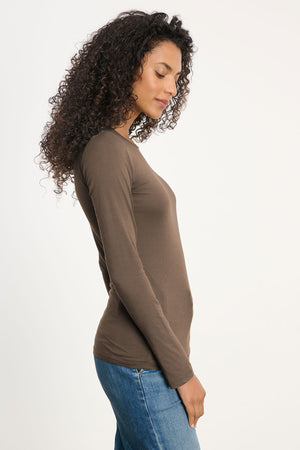 A person with curly hair wears the ZOFINA TEE by Velvet by Graham & Spencer— a fitted long sleeve crewneck tee—paired with blue jeans, standing in profile against a plain light background.