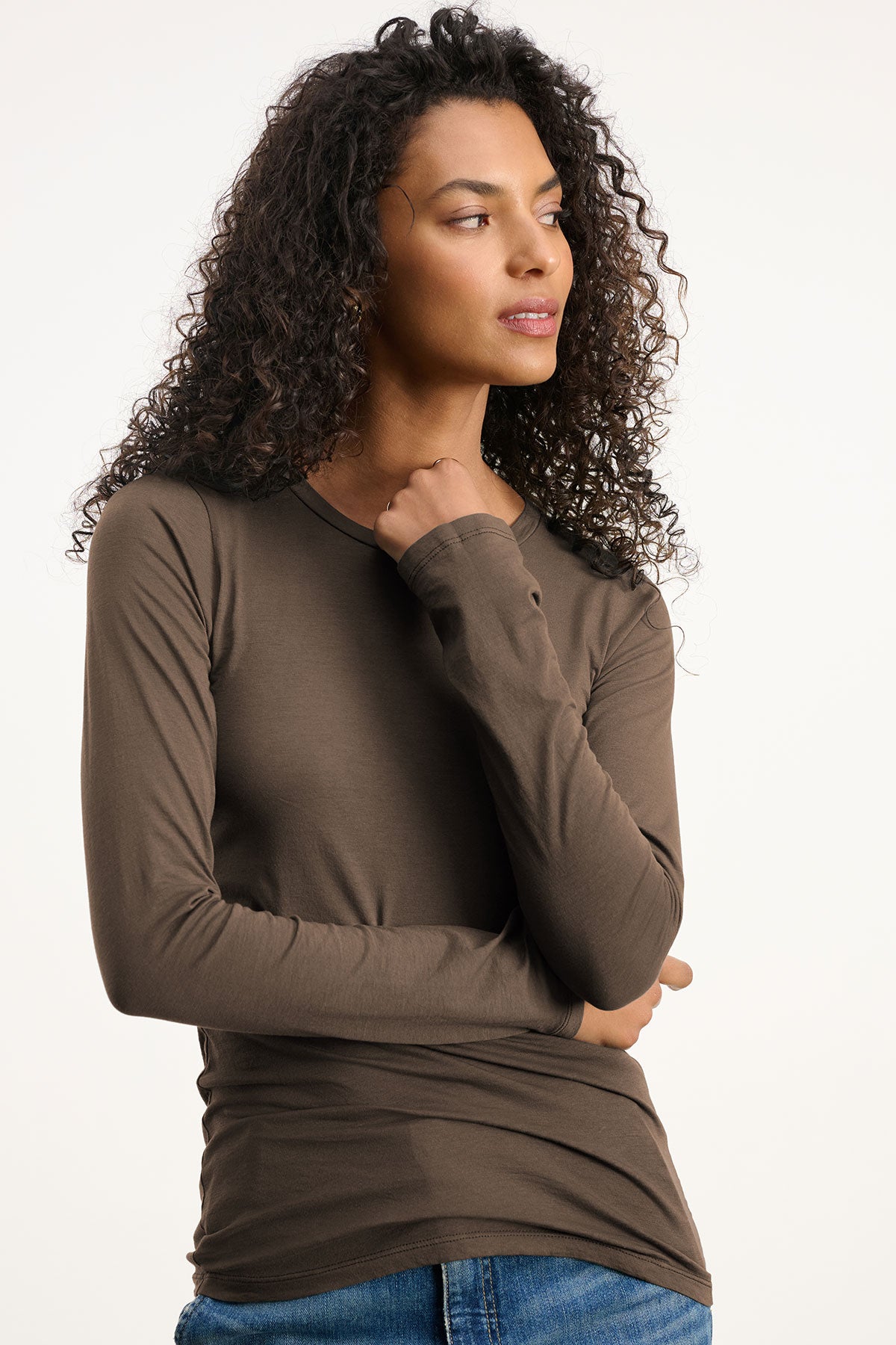 A woman with curly hair wears the ZOFINA TEE by Velvet by Graham & Spencer—a fitted, ultra-soft long sleeve with a classic crew neckline—in brown, paired with blue jeans. She stands against a plain light background, looking to the side.-39840187482305