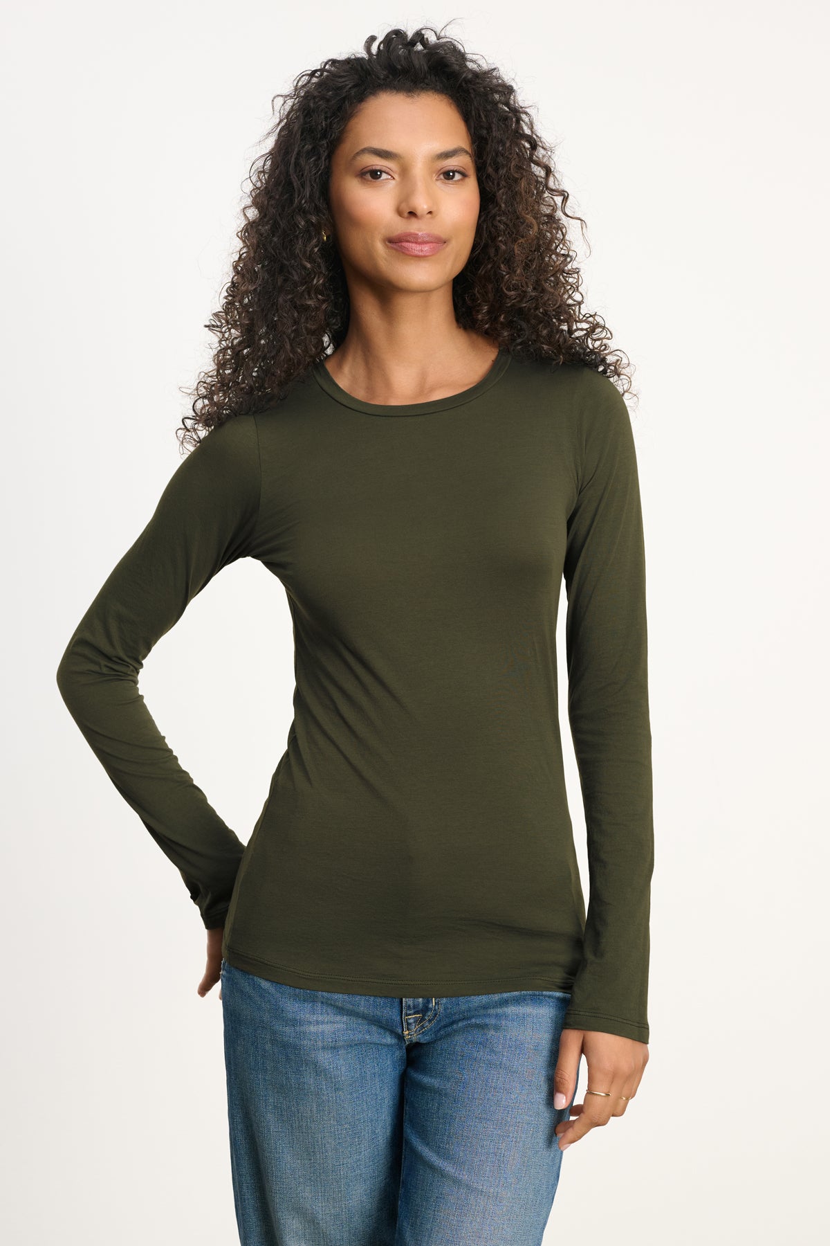 A woman with curly hair wears the Velvet by Graham & Spencer ZOFINA TEE in olive green, featuring a fitted silhouette and classic crew neckline, paired with blue jeans against a plain light background.