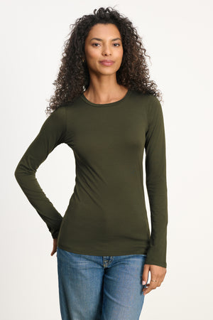 A woman with curly hair wears the Velvet by Graham & Spencer ZOFINA TEE in olive green, featuring a fitted silhouette and classic crew neckline, paired with blue jeans against a plain light background.