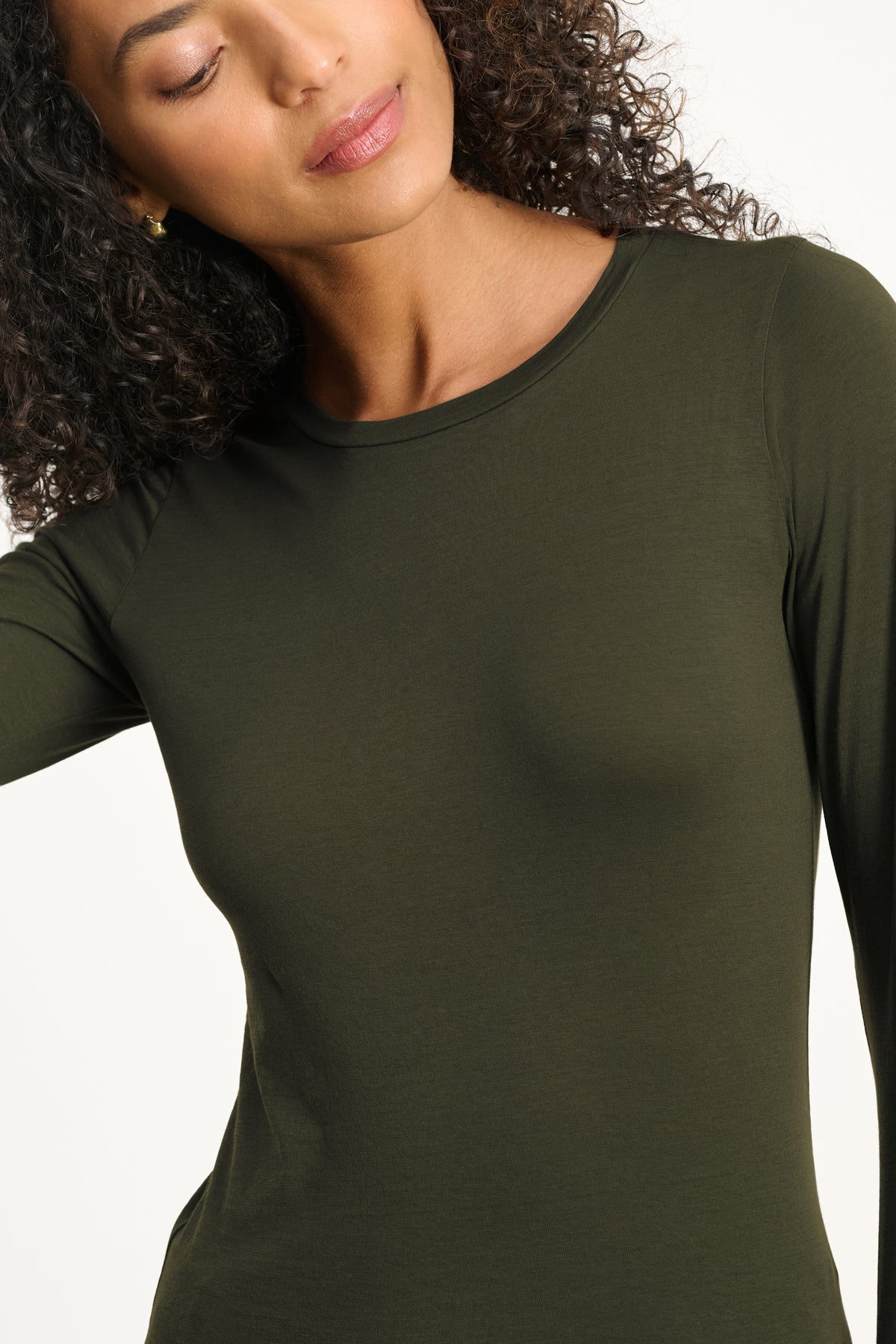 A person with curly hair wears the ZOFINA TEE by Velvet by Graham & Spencer in olive green, featuring a fitted long sleeve design, classic crew neckline, and ultra-soft gauzy whisper fabric, looking slightly downward.-40182503768257