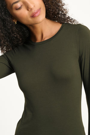 A person with curly hair wears the ZOFINA TEE by Velvet by Graham & Spencer in olive green, featuring a fitted long sleeve design, classic crew neckline, and ultra-soft gauzy whisper fabric, looking slightly downward.