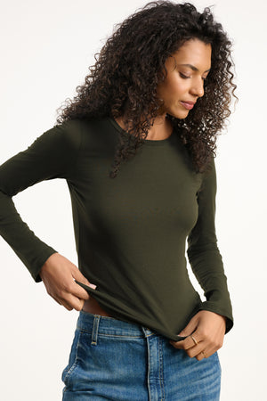 A woman with curly hair wears the Velvet by Graham & Spencer ZOFINA TEE, a fitted, ultra-soft long sleeve crewneck, paired with blue jeans as she looks down and holds the hem of her shirt against a plain background.