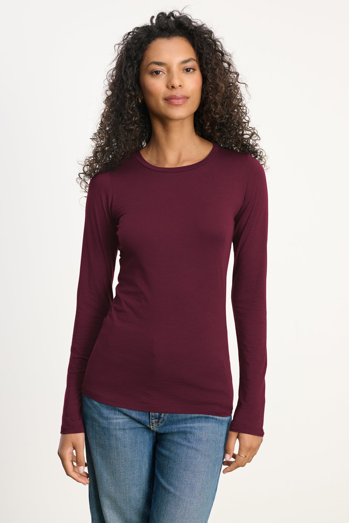 A person with curly hair wears the ZOFINA TEE by Velvet by Graham & Spencer—a fitted long sleeve burgundy crewneck—paired with blue jeans, standing against a plain light background.