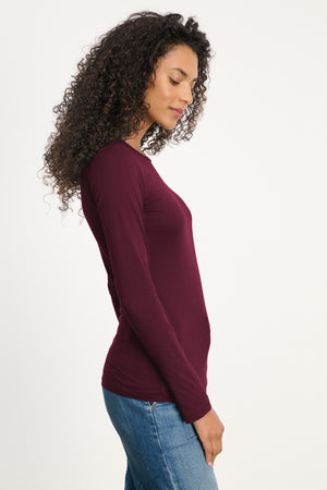 A person with curly hair stands sideways against a white background, wearing the ZOFINA TEE by Velvet by Graham & Spencer—a fitted long sleeve maroon crewneck tee—paired with blue jeans.