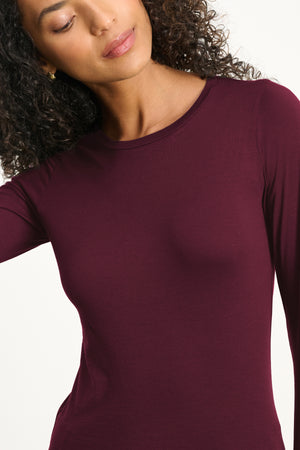 A woman with curly hair poses against a plain light background wearing the ZOFINA TEE by Velvet by Graham & Spencer, a fitted long sleeve top with a classic crew neckline.