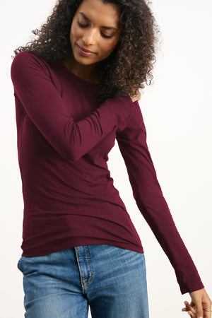 Someone wearing the ZOFINA TEE by Velvet by Graham & Spencer, an ultra-soft fitted long sleeve from the basics collection, with blue jeans, stands against a plain light background, looking down and touching their hair.