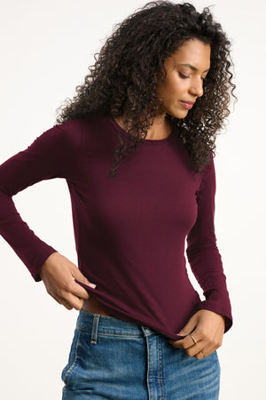 A woman in the ZOFINA TEE by Velvet by Graham & Spencer and blue jeans stands against a plain background, looking down as she adjusts her shirt hem.