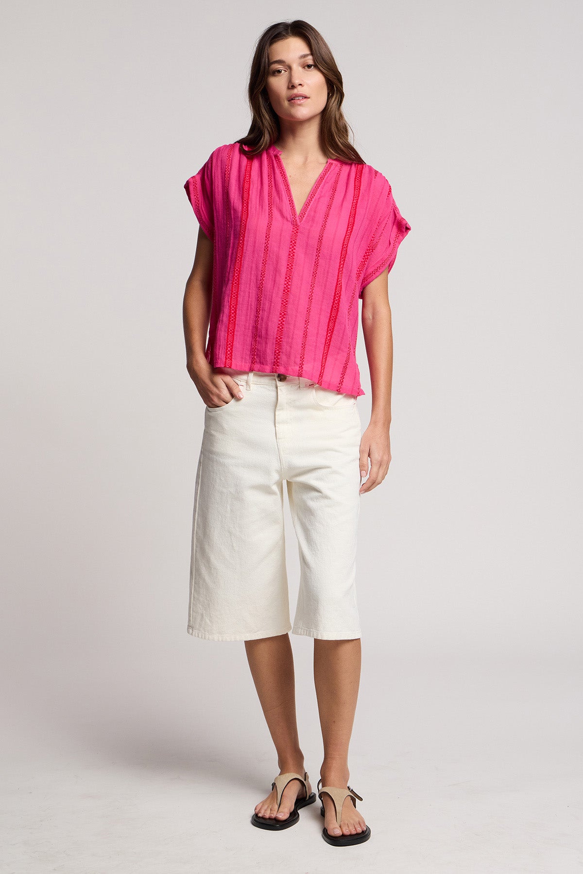 A woman stands facing forward against a plain light background, wearing the TALLULAH TOP in bright pink by Velvet by Graham & Spencer, paired with white knee-length shorts and black sandals.-41372222390465