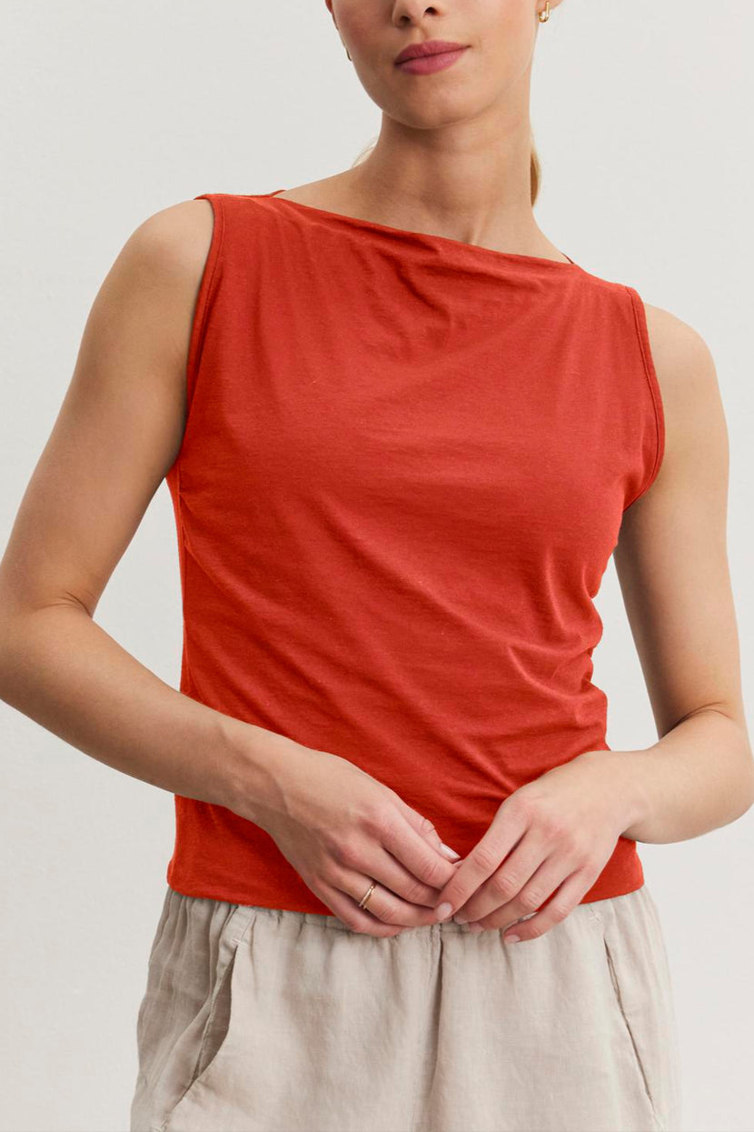A woman with blonde hair tied back, wearing a sleeveless red top and light beige pants, stands against a plain white background. She looks directly at the camera with a neutral expression, showcasing the EMILIA TANK TOP from Velvet by Graham & Spencer in modern minimal design made from soft cotton.