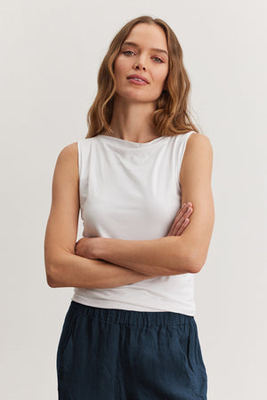 A woman with long wavy hair stands against a plain light background, wearing the ultra soft EMILIA TANK TOP from Velvet by Graham & Spencer and dark pants, arms crossed, with a neutral expression.
