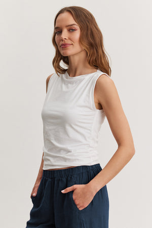 A woman stands with her hands in her pockets against a plain light background, wearing the EMILIA TANK TOP by Velvet by Graham & Spencer and dark blue pants.