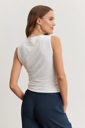 A woman stands facing away against a neutral background, wearing the EMILIA TANK TOP by Velvet by Graham & Spencer in white with dark blue pants; her left hand is in her pocket.