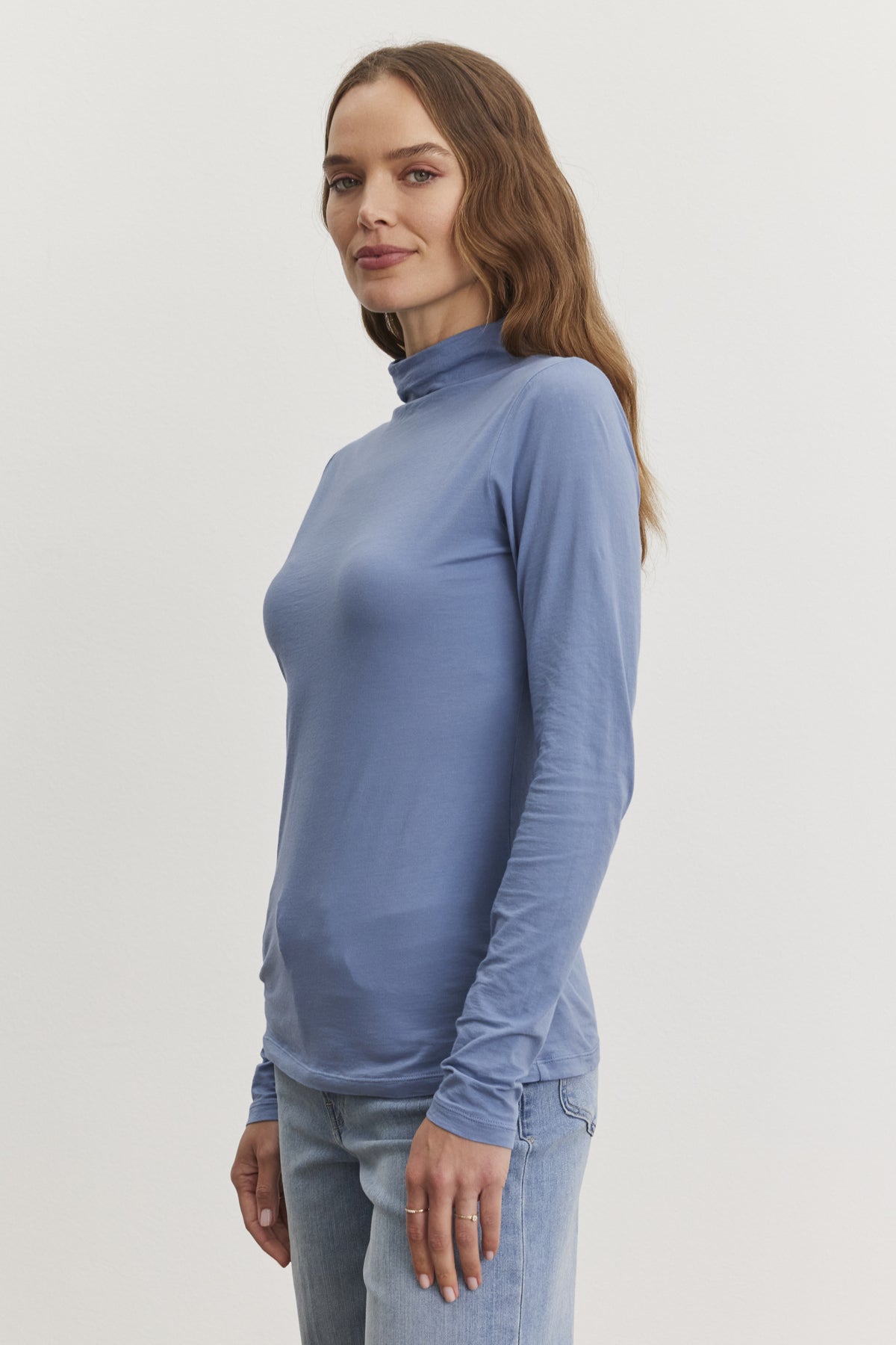 A person in a light blue TALISIA TEE by Velvet by Graham & Spencer and jeans stands against a plain background, showcasing this versatile wardrobe staple in contemporary fashion.