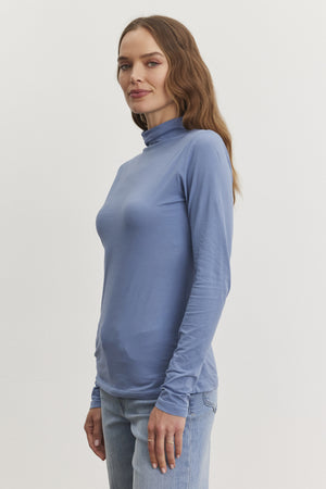 A person in a light blue TALISIA TEE by Velvet by Graham & Spencer and jeans stands against a plain background, showcasing this versatile wardrobe staple in contemporary fashion.