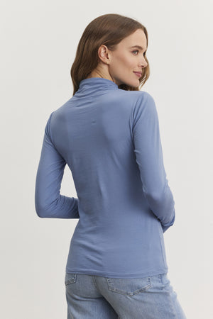 A woman wearing Velvet by Graham & Spencer's TALISIA TEE in blue long-sleeve mock neck and jeans, facing away from the camera against a plain background, exemplifies the stylish versatility of her wardrobe.